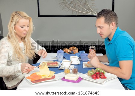 Attractive young couple enjoying breakfast with a healthy spread of coffee, cheese, cold meat, fresh fruit and rolls as they sit at a table indoors