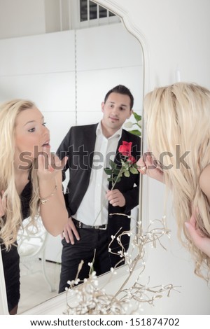 Romantic man holding a single long stemmed red rose watching a beautiful young blond woman applying her makeup in the mirror