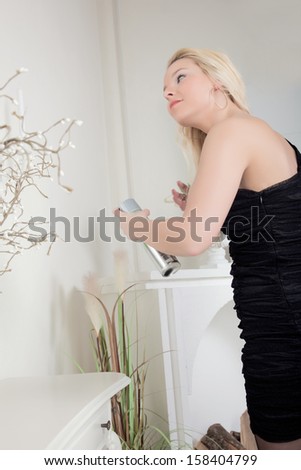 Woman spraying her long blond hair with hairspray as she dresses for a night out in a stylish black dress