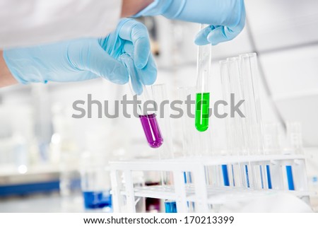 The Chemist takes up two test tubes with a  purple and green liquid.