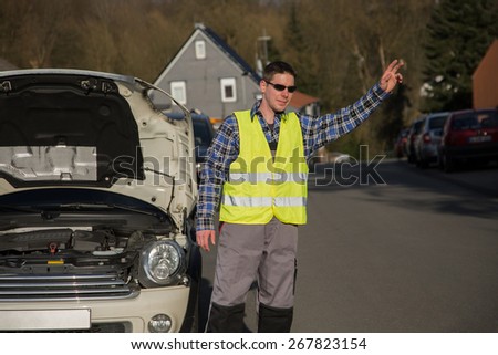 A young man dressed with an reflective vest is standing beside his car and ask for help with his hand.