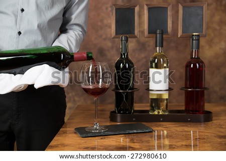 A Waiter is pouring red wine into a glass with three bottles of wine in the background.