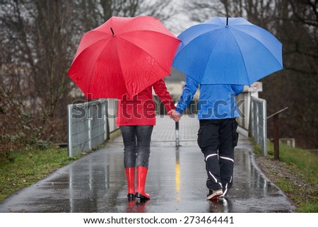 A young couple is walking hand in hand in the rain through a park.
