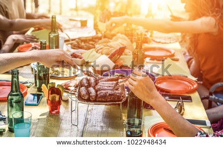 Group of happy friends eating and drinking beers at barbecue dinner on sunset time - Adult people having meal together outdoor - Focus on fork sausages - Summer lifestyle, food and friendship concept
