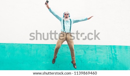 Senior crazy man jumping and listening music outdoor - Happy mature male celebrating and dancing outside - Joyful elderly lifestyle concept - Focus on him