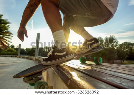 Silhouette of young man performing with skateboard up wood bench at sunset in urban city park - Skater having fun with back sunlight - Extreme sport concept - Focus on left shoe - Warm filter