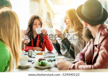 Young friends toasting coffee and doing breakfast in bar bakery shop - Happy hipster people drinking cappuccino and eating muffins - Friendship concept - Focus on center girl - Warm filter