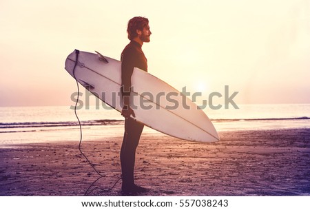 Man surfer carrying his surfboard at sunrise - Hipster male in wetsuit waiting for the high waves on beach - Extreme sport concept - Focus on male silhouette - Matte filter with soft blue vignette