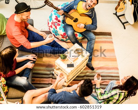 Group of trendy friends having fun in hostel living room - Happy young backpackers enjoying time together playing music with guitar and drinking beer - Focus on musician and wood table - Warm filter
