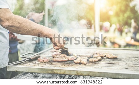 Man cooking meat at dinner barbecue - Chef grilling meat in park outdoor - Concept of eating bbq outdoor during summer time - Focus on left man hand - Warm vintage filter with back sunlight