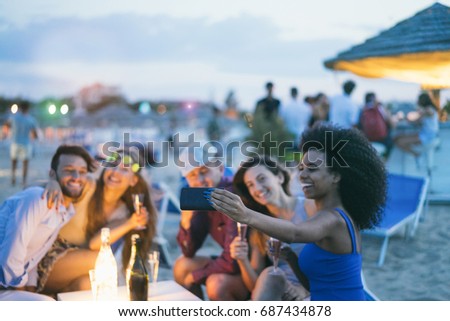 Happy friends taking selfie with smartphone at beach party outdoor - Young people having fun together at kiosk bar drinking champagne  - Focus on african girl hand phone- Youth and summer concept