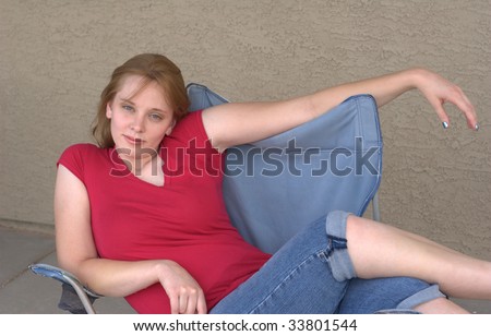 A gorgeous young woman sits on a blue camping chair leaning to a side looking at the viewer in a flirty manner