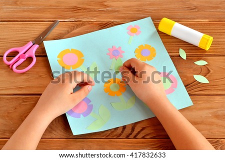 Child doing a card with flowers. Child holds a paper leaf in his hands and sticks it. Glue, scissors, greeting card on a wooden table. Paper flower craft for kids. Child art project.
