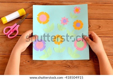 Child holds a card with flowers in his hands. Glue, scissors on a wooden table. Paper flower craft for kids. Child art project.