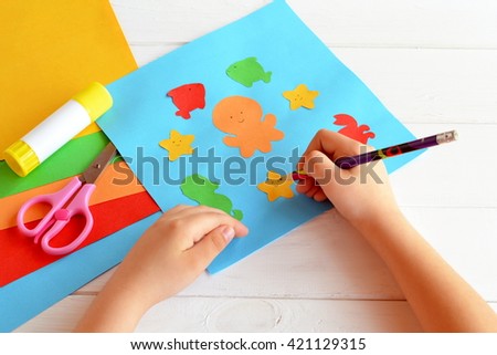 Child holds a pencil and draws. Child doing a card with sea animals and fish. Sheets of colored paper, scissors, glue, set for kids art