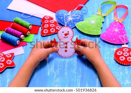 Child holds a felt snowman ornament in his hands. Christmas sewing crafts concept. Christmas tree, heart, star, snowman crafts, sewing kit, bright felt sheets on blue wooden background