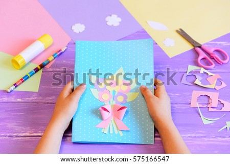 Small child made a greeting card with flowers for mom. Step. Child holds a card in his hands. Tools and materials for children\'s art creativity on table. Mother\'s day or March 8 greeting card DIY idea