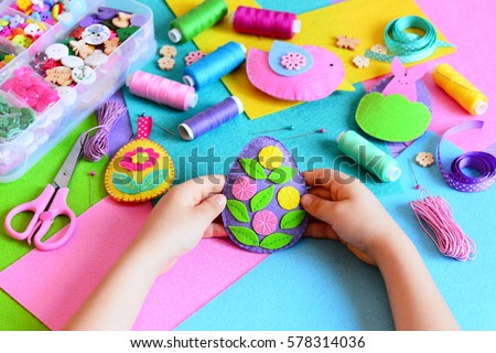 Child made a felt Easter egg ornament. Small child holds a felt Easter egg ornament in his hands. Easter crafts, craft tools and materials on a table. Festive spring crafts concept