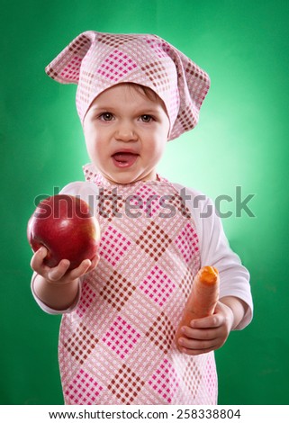 the baby girl with a kerchief and kitchen apron holding an vegetable isolated on the green background