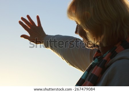A senior woman reaching out to the sun in the sky, as if waiting for someone or seeing someone just around the corner.