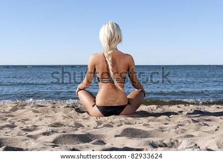 blonde girl in a black bathing suit sits with his back to the beach in the sand and meditate
