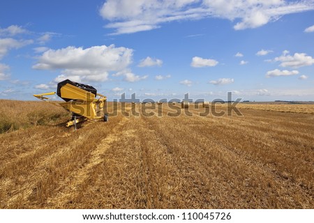 a yellow cutting machine parked in a stubble field  with round bales pattern line and texture next to a crop of ripe canola under a summer blue sky in the yorkshire wolds england