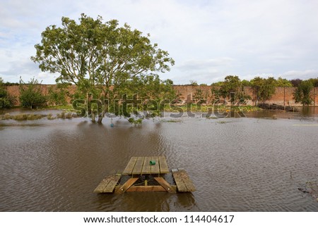 a flooded walled garden in autumn with a submerged picnic bench with trees and vegetation under a cloudy sky