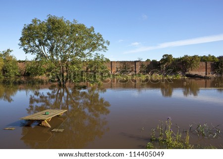 a flooded walled garden in autumn with submerged picnic bench and trees reflected in the water under a clear blue sky