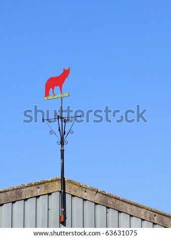 a weather vane in the shape of a red fox on a farm building under a blue sky