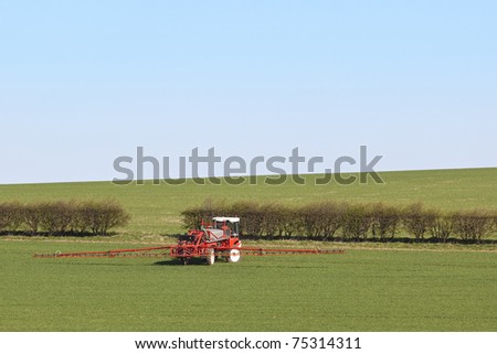 an english landscape with a red and white crop sprayer working in a field of cereals under a clear blue sky in springtime