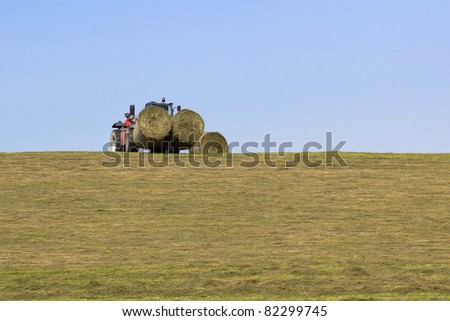 a red farm loader collecting big round hay bales on a grassy hillside under a blue sky