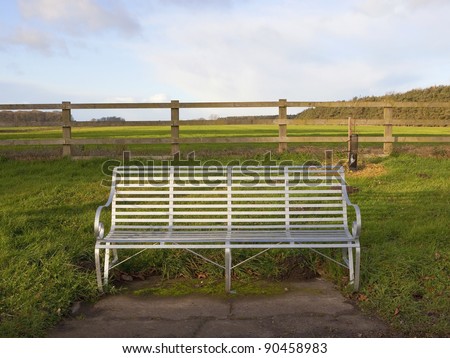 a rural landscape with a silver metal bench rustic wooden fence with fields and woods behind