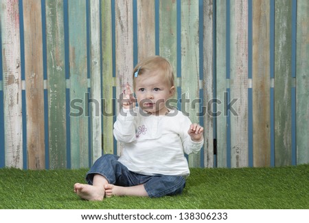 baby seated on grass in front of a weathered fence saying no