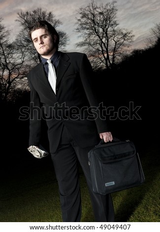 Business man standing in a suit, holding a brief/laptop case and a newspaper.