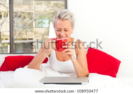 Portrait of a pretty woman sitting at her bed with a laptop and cup of coffee or tea looking happy