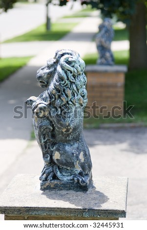 A pair of decorative lions atop pedestals on either side of a driveway, with a sidewalk which runs through the scene.