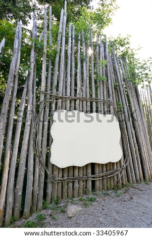 A blank sign / placard framed with wooden tree limbs, surrounded by logs, creating a long wall.