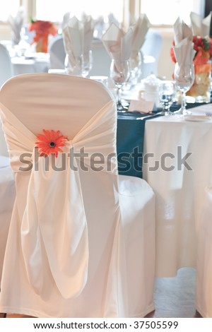 Focus on the back of a wedding chair and chair cover with ornamental orange daisy, and tables and bright window with natural daylight in the background.