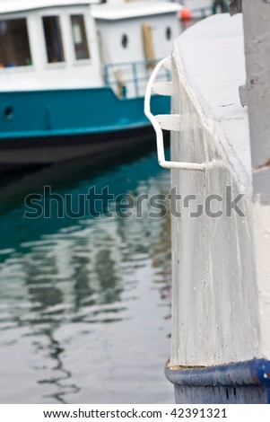 The corner of an old boat, showing the texture of it's painted hull, with another boat and reflections in the water behind.