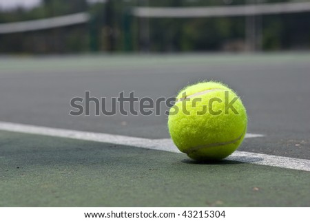A closeup of a yellow tennis ball just outside of the base line on an asphalt tennis court.