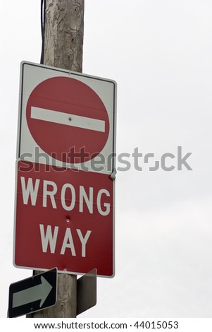 A wrong way sign on a hydro pole, with \'one way\' signs below indicating the direction of travel, against an overcast sky.