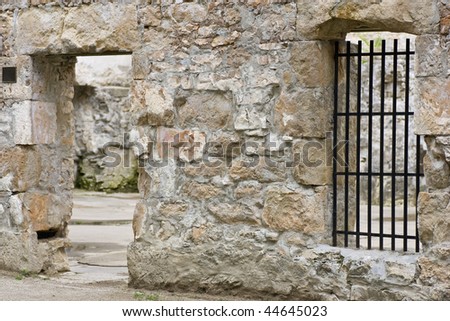 An old ruin wall outside, with an opening into another outdoor area, and window with bars on it.