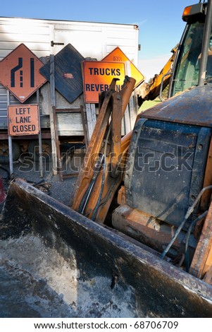 A closeup of a front loader construction vehicle, with a bunch of street and construction signs behind, resting on a portable trailer.