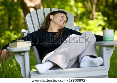 A woman sitting comfortably in an outdoor adirondack chair, with a coffee mug on one arm, and a book on the other.