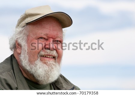 A closeup of a senior male with white beard and hair, in a baseball cap, isolated against sky.