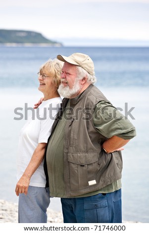 Two seniors on a rocky beach.  The husband has his around around his wife, as they both look away to camera left.