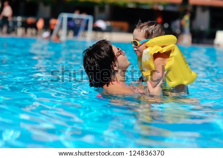 In the sunny day in the summer the boy in sun glasses and in a life jacket plays pool with mum