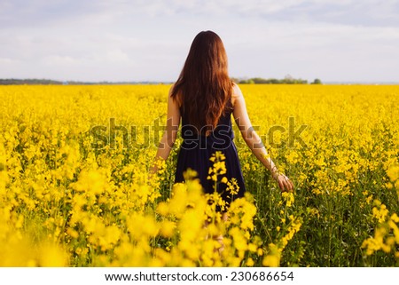 Young woman in sleeveless dress enjoying sunlight and nature on yellow blooming rapeseed field