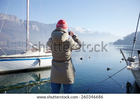 Rear view of tourist taking photo by smartphone of snowy mountain landscape and foggy lake with moored yachts near berth
