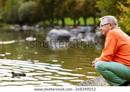 Profile of aged man in glasses sitting near pond in park watching ducks